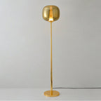 Gold Clérachic floor lamp lit warm, its gilt glass head glowing, standing on a round gold base against grey.