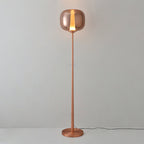A rose gold Clérachic floor lamp stands lit on a round base, its thin stem rising to the glass head against a pale wall.