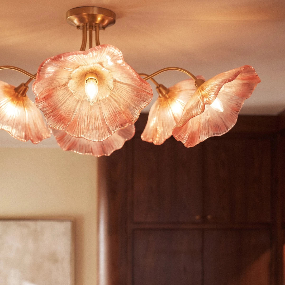 Clérachic ceiling light with blush-red ribbed glass flower shades on brass arms, glowing above a dark wood cabinet.