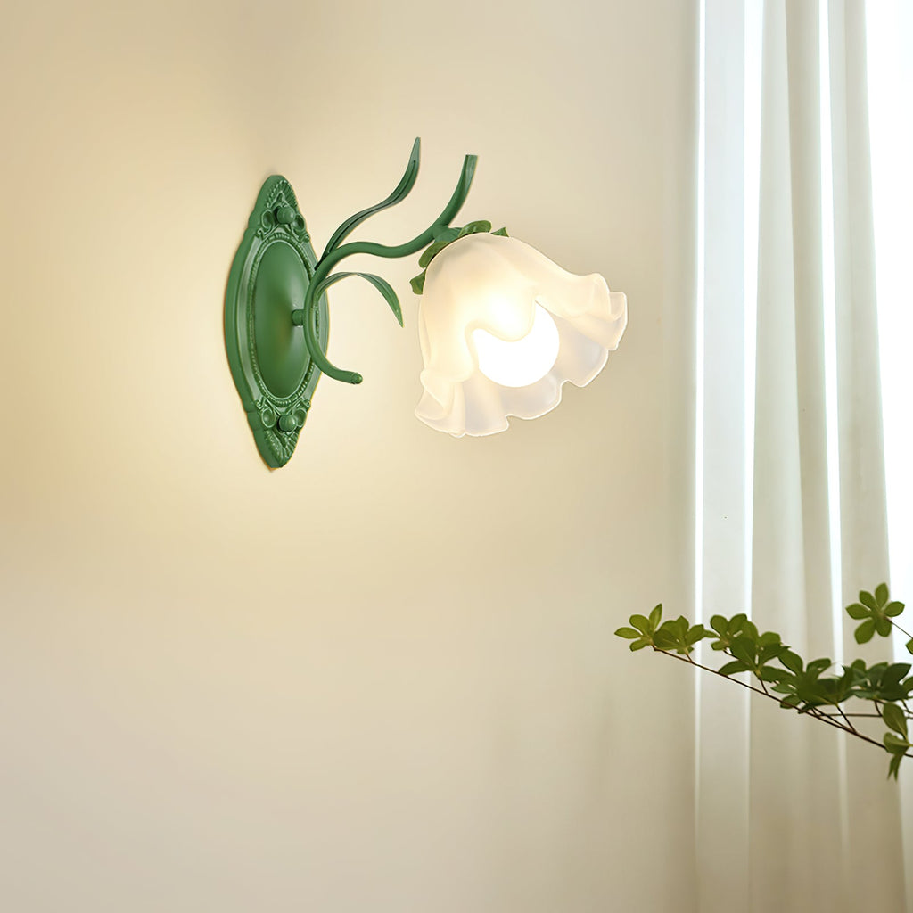 Lit single-flower Clérachic wall light in green, casting warm light on a cream wall beside a sheer curtain.