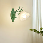 Lit single-flower Clérachic wall light in green, casting warm light on a cream wall beside a sheer curtain.