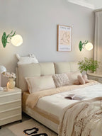 Two green Clérachic wall lights flank a cream bedroom bed, one on each side of a framed print above the bedside tables.
