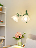 A Clérachic wall light with two ruffled flower shades lit above a shelf, a pink hydrangea and a sofa arm below.