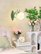 Single green Clérachic wall light with a round opal glass shade, above a white bedside table with pink roses.