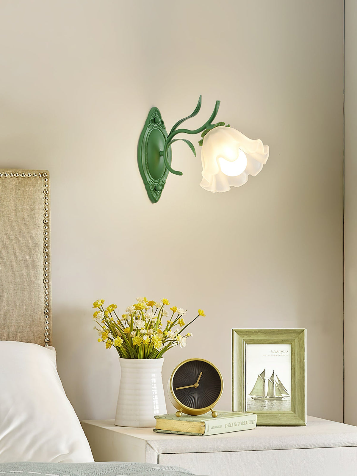 Green Clérachic wall light with a ruffled glass flower shade, above a bedside table with yellow flowers and a clock.
