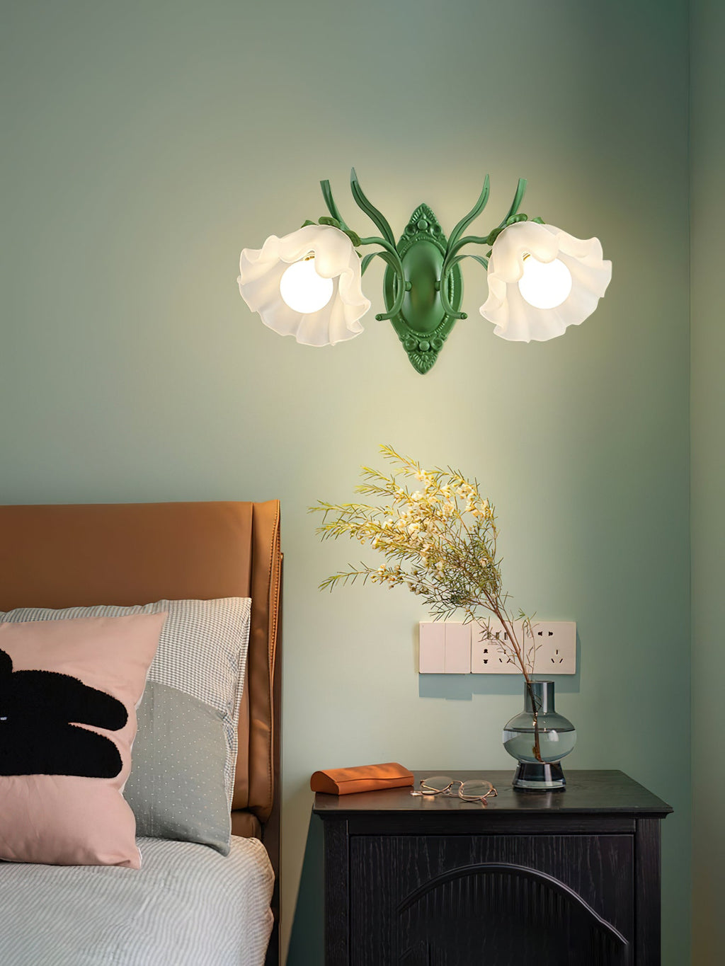 Two lit flower shades on a green Clérachic wall light, set against a sage wall above a dark bedside cabinet.