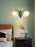 Two lit flower shades on a green Clérachic wall light, set against a sage wall above a dark bedside cabinet.