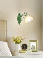 A single green Clérachic wall light with a frosted flower shade over a headboard, yellow flowers and a framed print beside it.