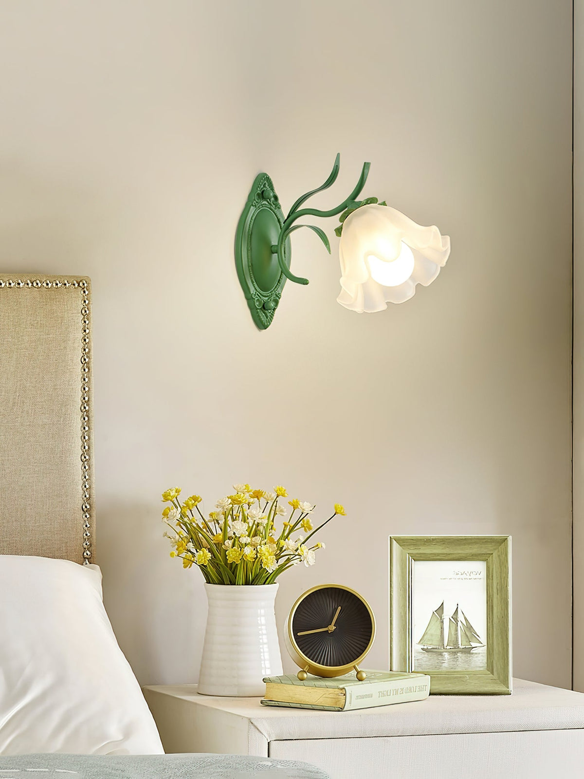 A single green Clérachic wall light with a frosted flower shade over a headboard, yellow flowers and a framed print beside it.