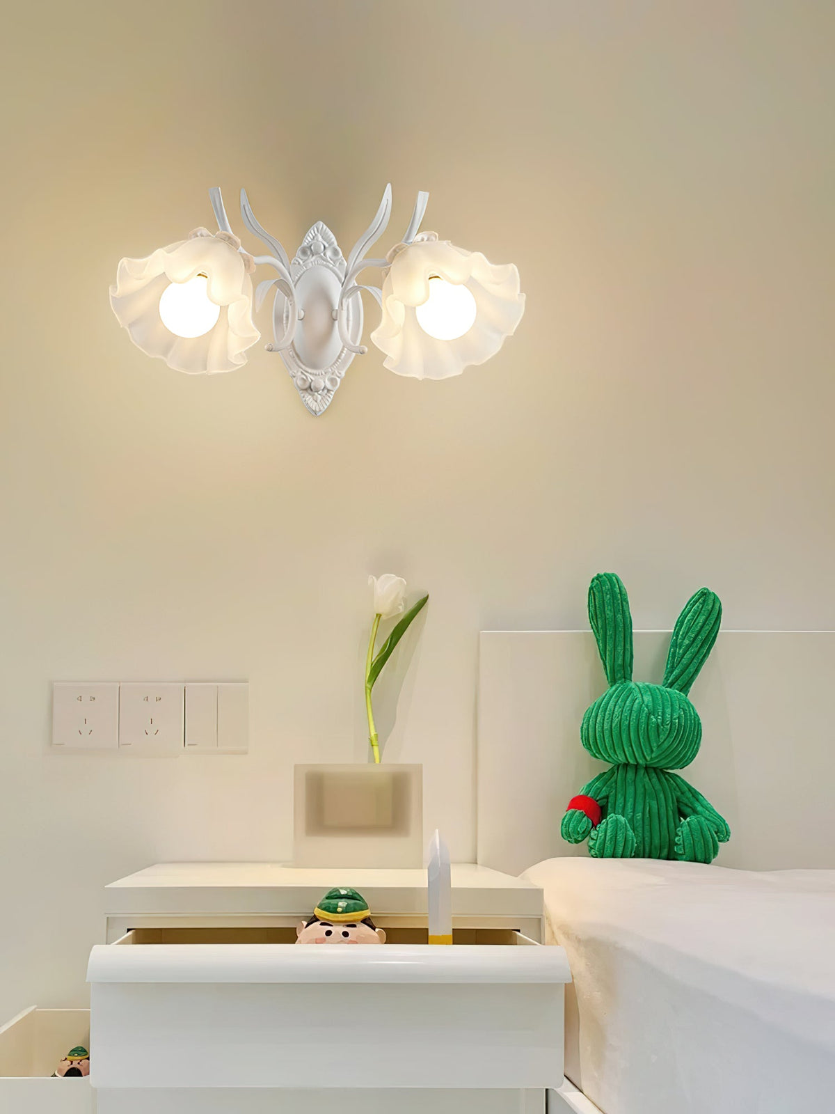 A white Clérachic double wall light with two ruffled flower shades, above a white bedside cabinet and a green toy rabbit.