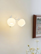 White Clérachic double wall light with two round opal glass shades, glowing on a cream wall beside a wooden frame.
