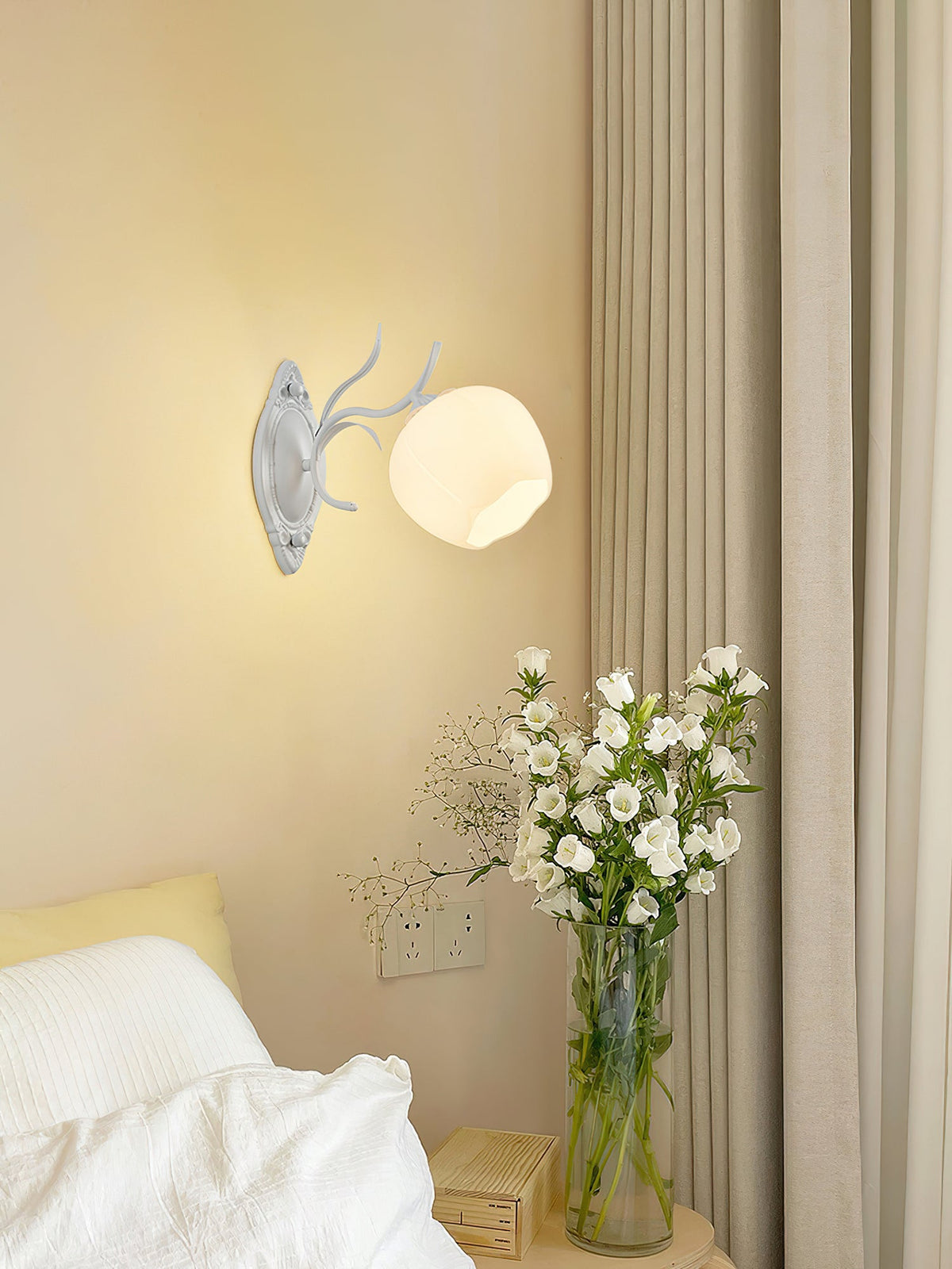 A white Clérachic wall light with a round opal shade, lit beside a beige curtain and a vase of white flowers.