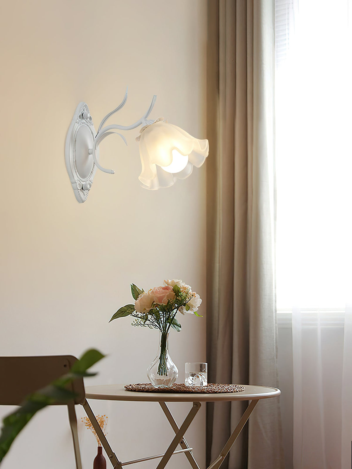 White Clérachic wall light with a ruffled flower shade, lit beside a curtain and a small table with pink flowers.