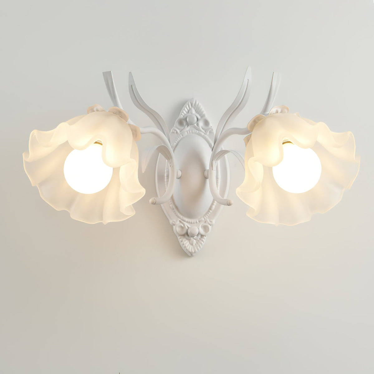 A white Clérachic double wall light, lit, with two ruffled flower shades and a carved backplate on a grey wall.