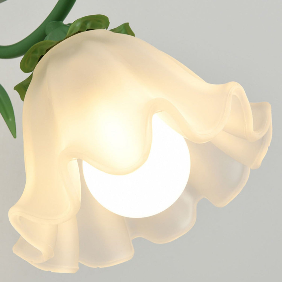 Close view of the lit, frilled glass flower shade on a green Clérachic wall light, with leaf details on the arm.