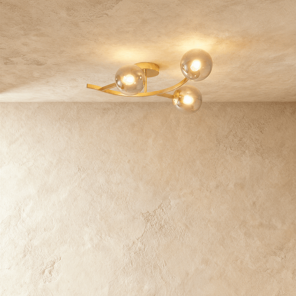 Clérachic three-globe ceiling light on a curved gold arm, smoked glass globes lit warm against limewash plaster.