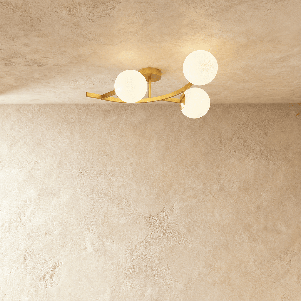 Clérachic three-globe ceiling light on a curved gold arm with opal white glass globes, lit against plaster.