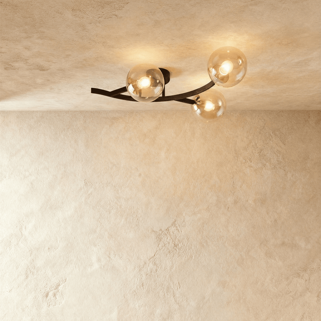 Clérachic three-globe ceiling light on a curved black arm, amber-tinted clear glass globes lit warm on plaster.
