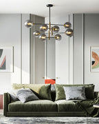Distant view of a Clérachic chandelier with glass globes hanging above a deep green velvet sofa in a pale grey room with framed prints.