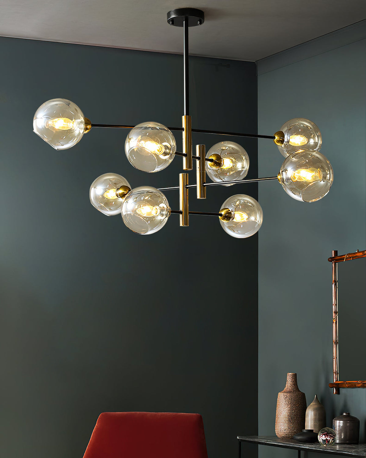 Lit amber globes on a Clérachic chandelier in a dark green room, with a red chair, ceramic vases and a wall mirror below.