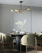 Clérachic chandelier with small amber globes above an oval dining table with olive green chairs and a vase of pampas grass.