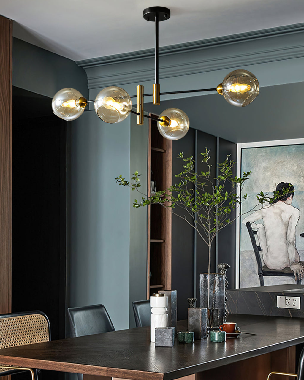 Clérachic chandelier with amber globes over a dark dining table, in a blue-grey room with a figure painting and a tall leafy stem.