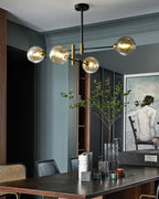Clérachic chandelier with amber globes over a dark dining table, in a blue-grey room with a figure painting and a tall leafy stem.