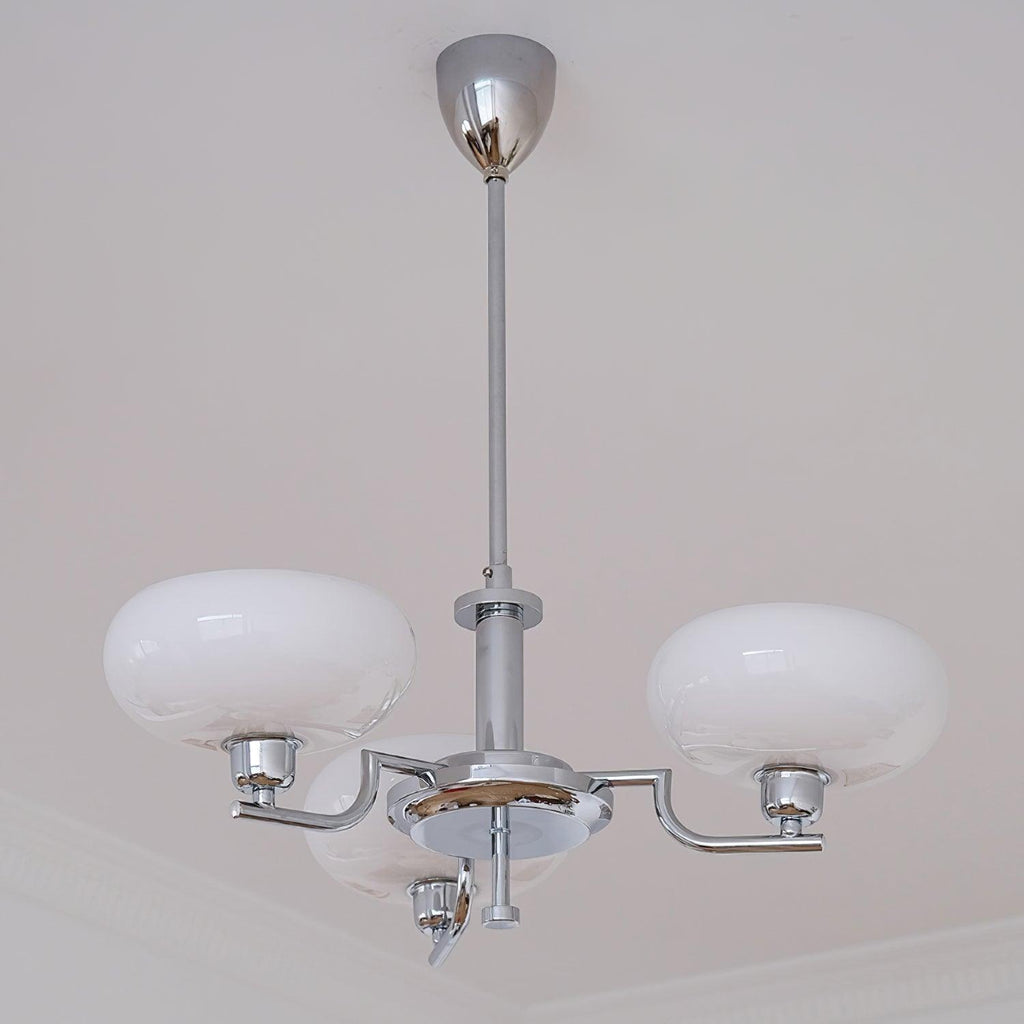 Clérachic three-head chrome chandelier with milk-glass globes, low-angle view beside crown moulding.