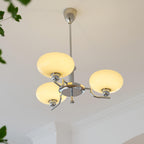 Clérachic three-head chrome chandelier lit warm, cream globes glowing above a plant and crown moulding.