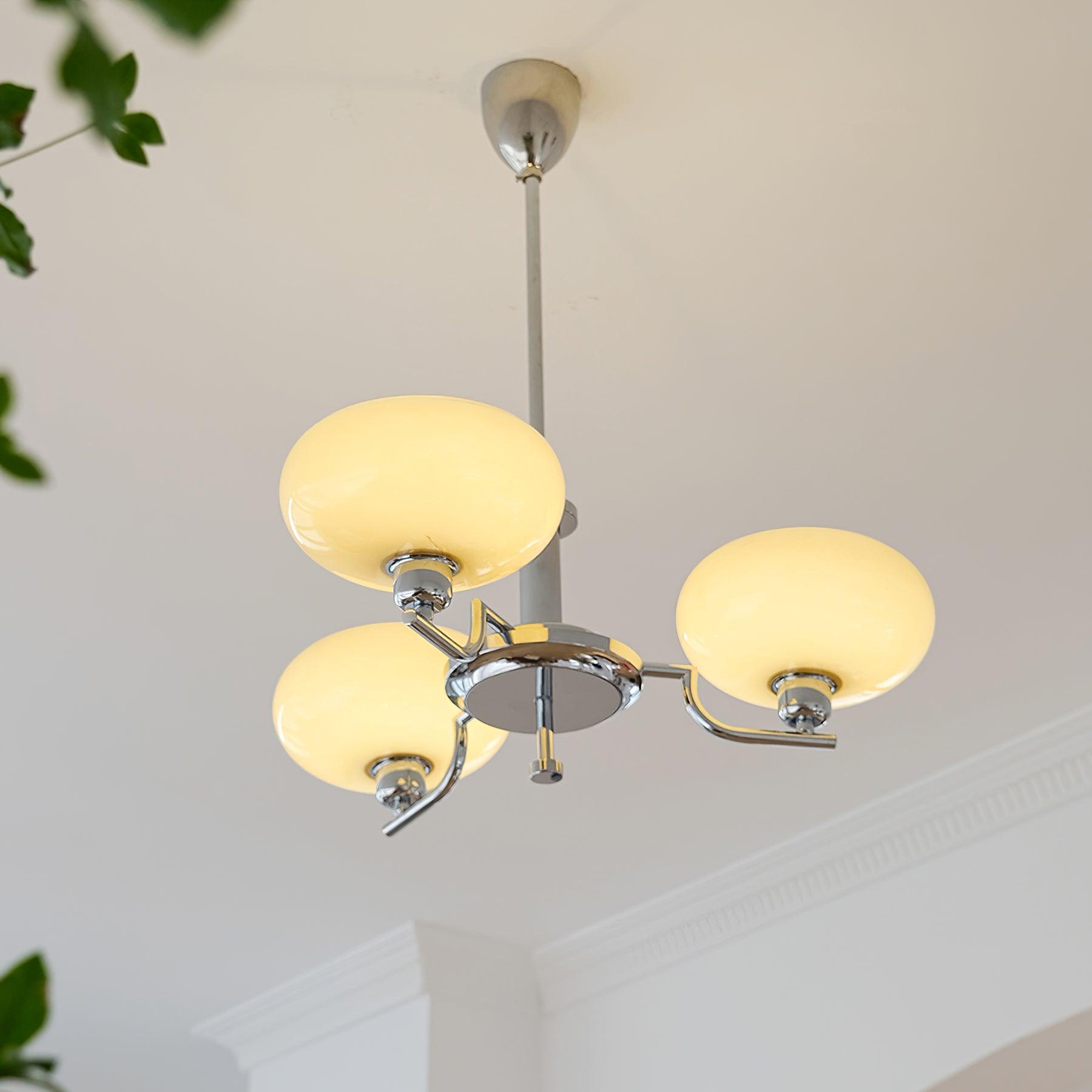 Clérachic three-head chrome chandelier lit warm, cream globes glowing above a plant and crown moulding.