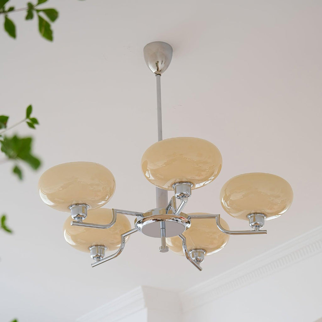 Clérachic five-head chrome chandelier with cream globes, houseplant leaves framing a white ceiling.