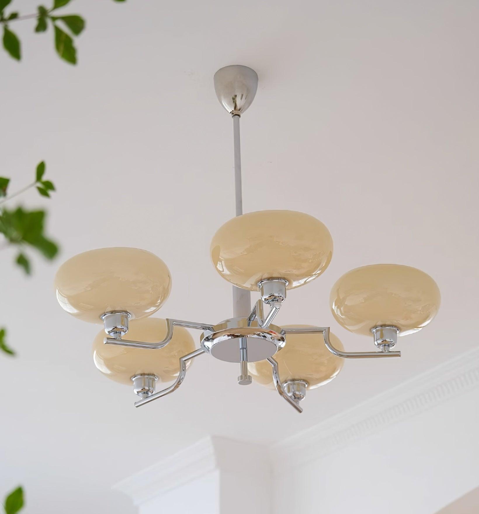 Clérachic five-head chrome chandelier with cream globes and a small hub finial, plant leaves at the left.