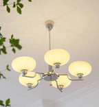 Clérachic five-head chrome chandelier lit warm, cream globes glowing above a plant and crown moulding.