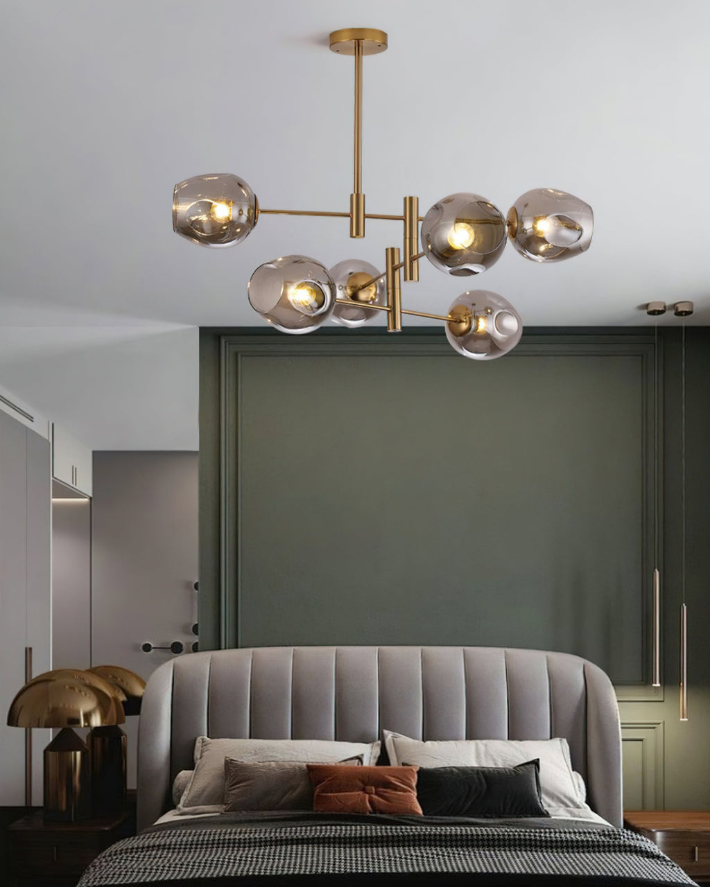 Smoky grey globes lit on this Clérachic gold chandelier over a bed with a grey channelled headboard and green-grey walls.