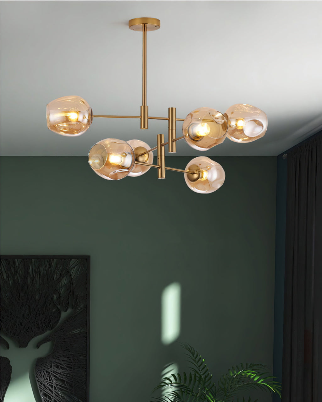 Amber globes on a gold Clérachic chandelier against a deep green wall, with a dark curtain and a stag print below.