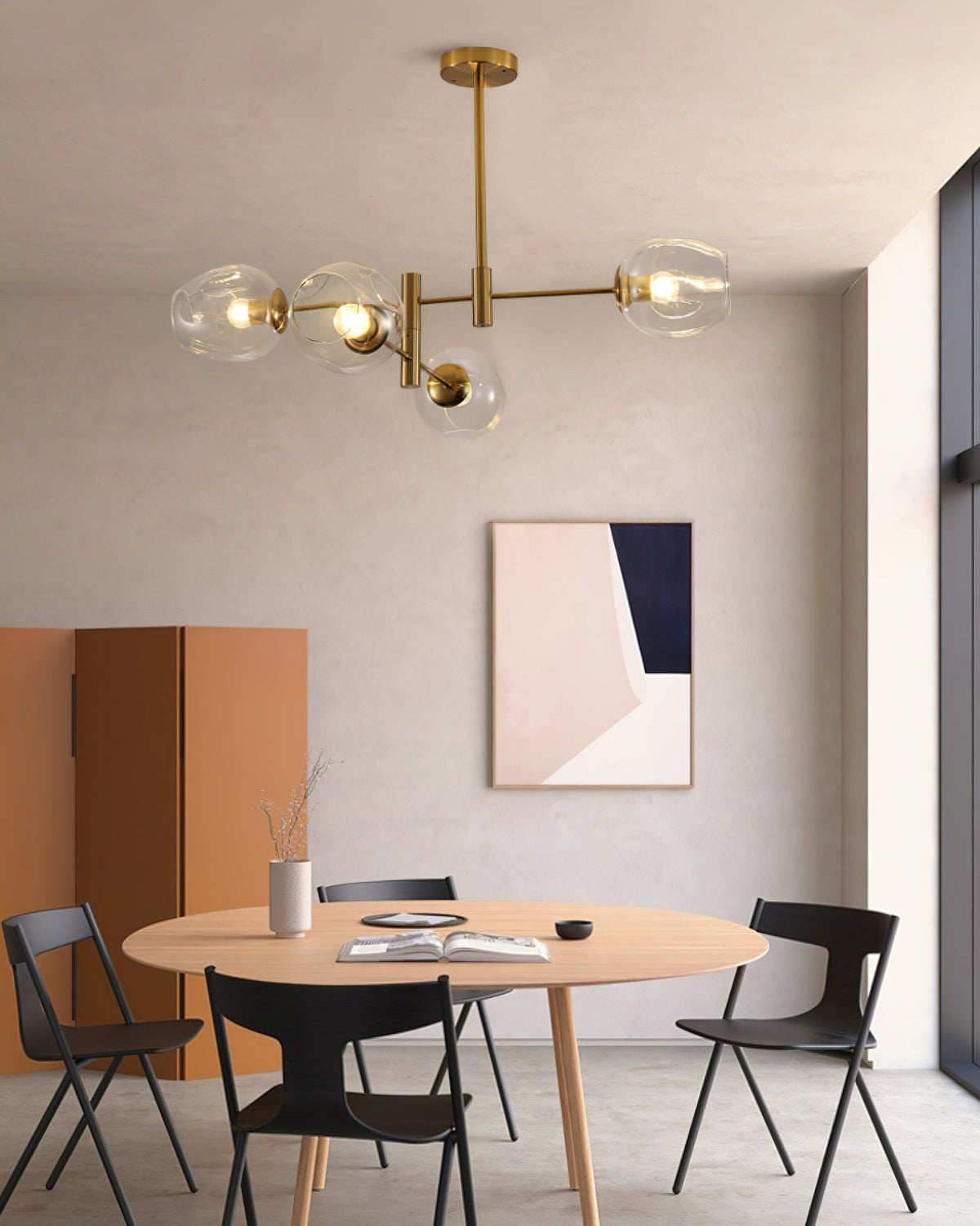 Four clear glass globes on a gold-finish Clérachic chandelier above a round table with black chairs and an orange folding screen.