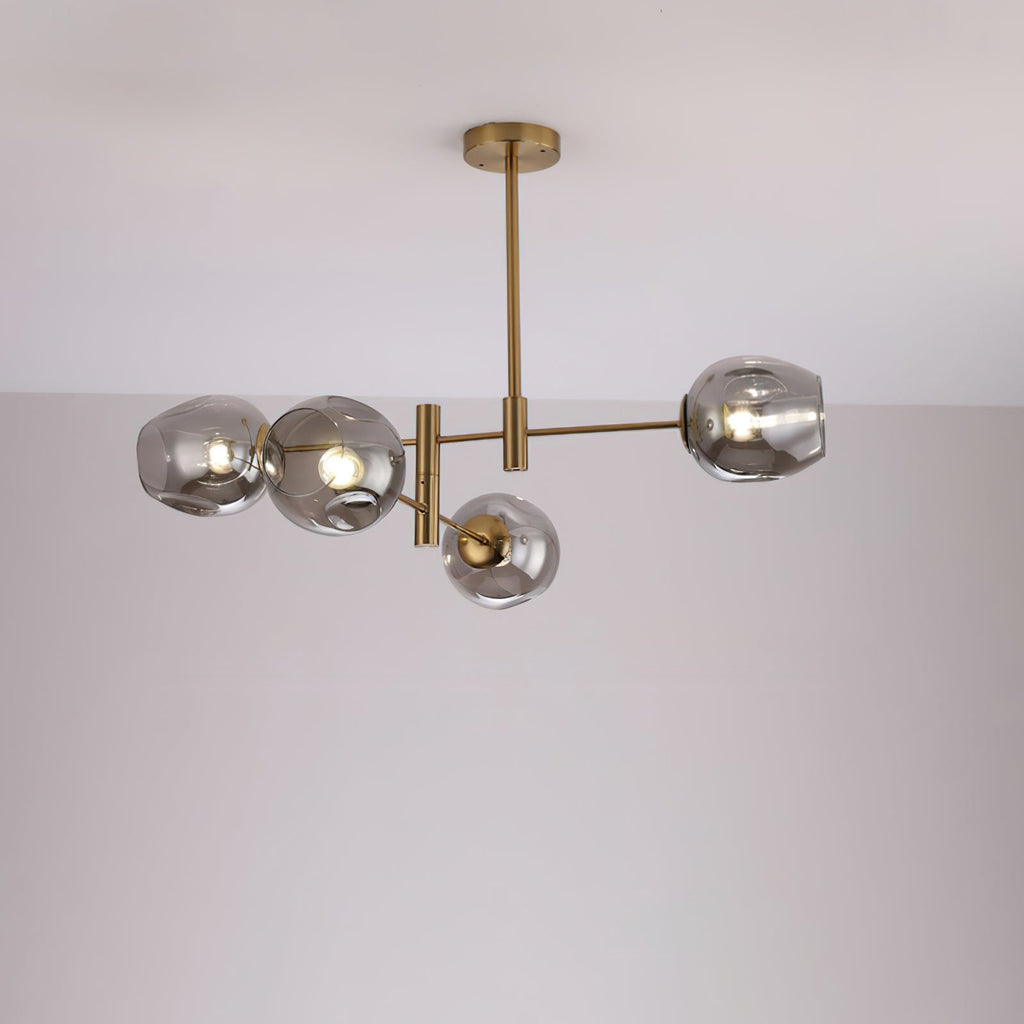 Four smoky grey glass globes lit on a gold-finish Clérachic chandelier with a slim rod, against a pale wall.