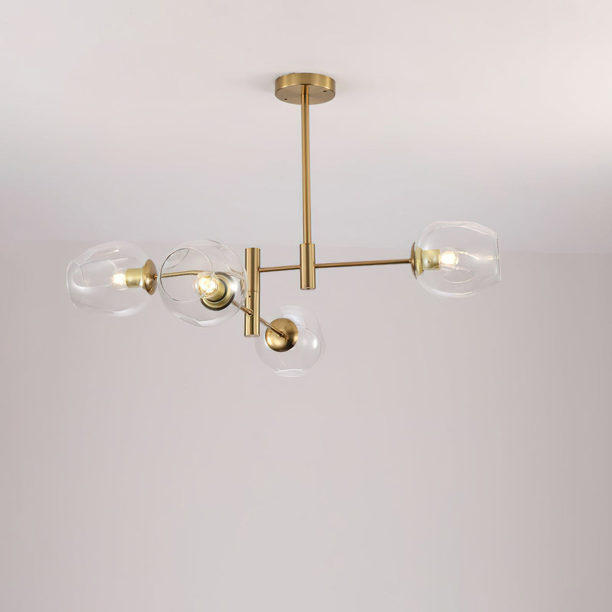 Four clear glass globes lit on a gold-finish Clérachic chandelier with a slim ceiling rod, against a pale wall.