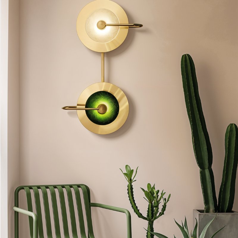 Clérachic two-head brass wall lamp with clear and green glass domes on a blush wall, beside a green chair and tall cacti.