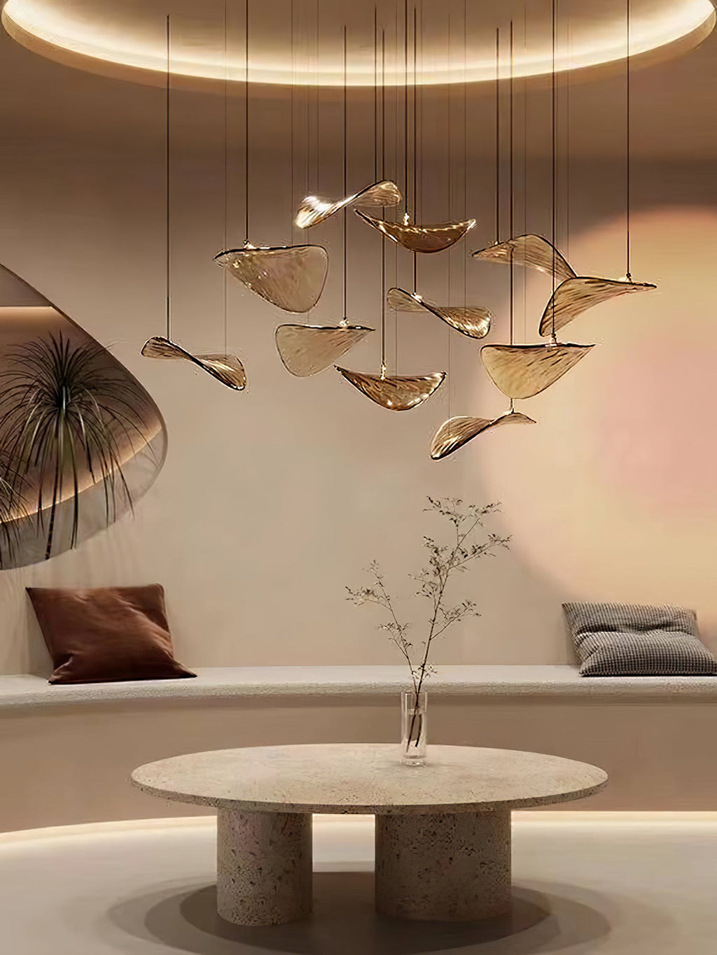 Round table with a bare twig vase sits under a Clérachic pendant of amber glass leaves, in a warm room with a curved bench.