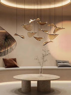 Round table with a bare twig vase sits under a Clérachic pendant of amber glass leaves, in a warm room with a curved bench.