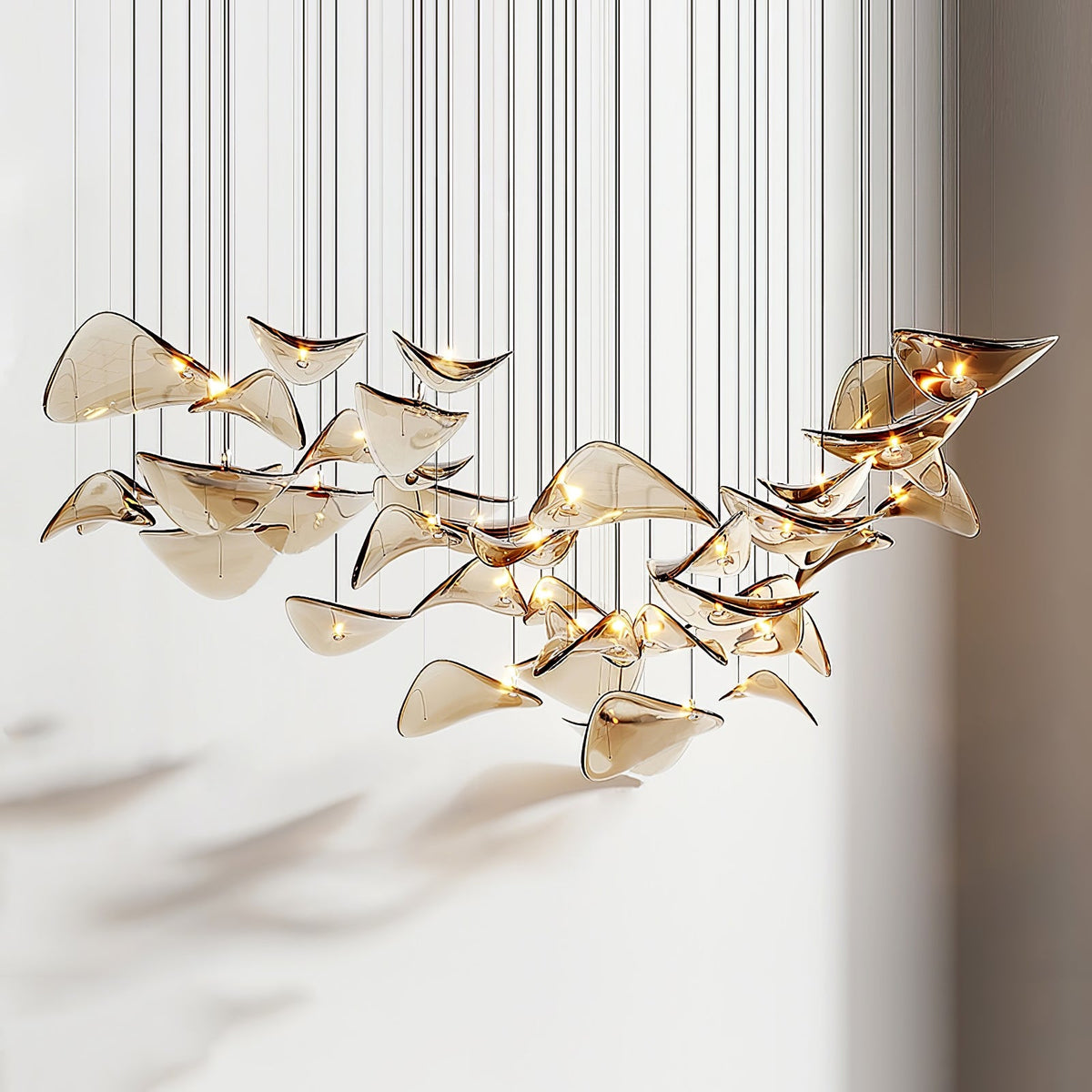 Wide arc of amber glass leaves on thin cables, lit from within, from this Clérachic pendant hung in front of a pale wall.