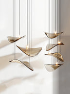 Six amber glass leaves hung in a loose group on fine cables, each with a small light, against a pale wall with leaf shadows from Clérachic.