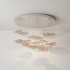 Large round chrome canopy over a hanging group of amber glass leaves, seen against a pale wall with soft window light, from Clérachic.
