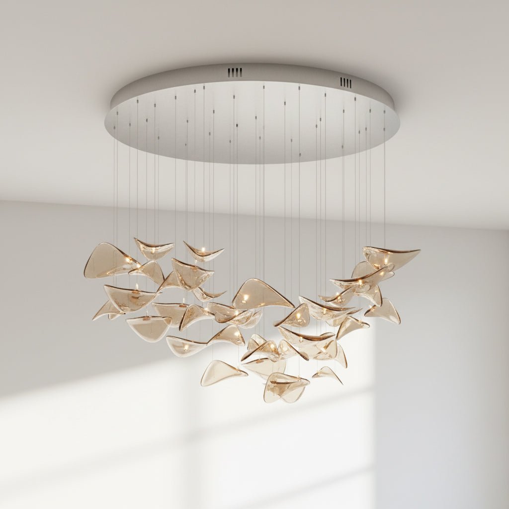 Round chrome canopy with fine cables and a loose group of amber glass leaves hung below it, from this Clérachic pendant, against a pale wall.