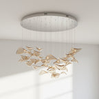 Round chrome canopy with fine cables and a loose group of amber glass leaves hung below it, from this Clérachic pendant, against a pale wall.