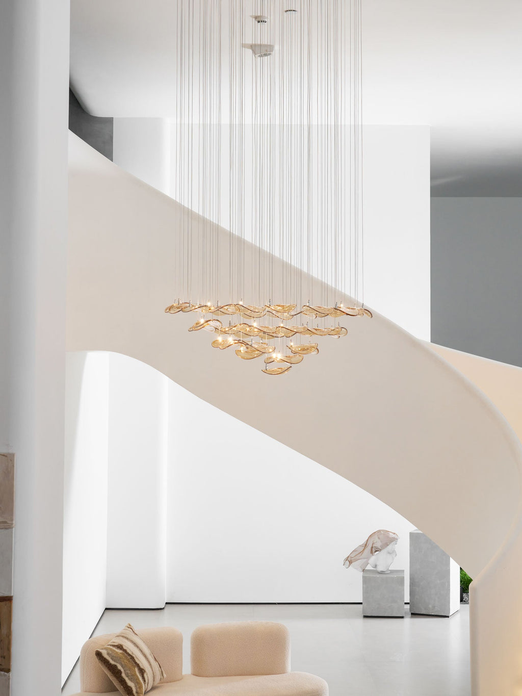 Tall white stairwell with a pale curving stair, where a Clérachic amber leaf pendant hangs high above a sofa and a plinth.