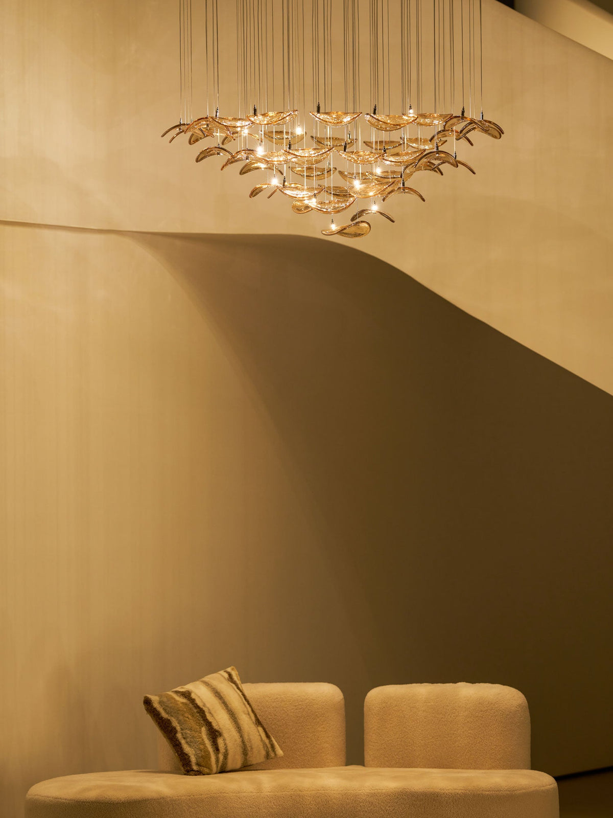 Dim room with a curved cream stair and a low sofa, with a Clérachic amber leaf pendant glowing high in the background.