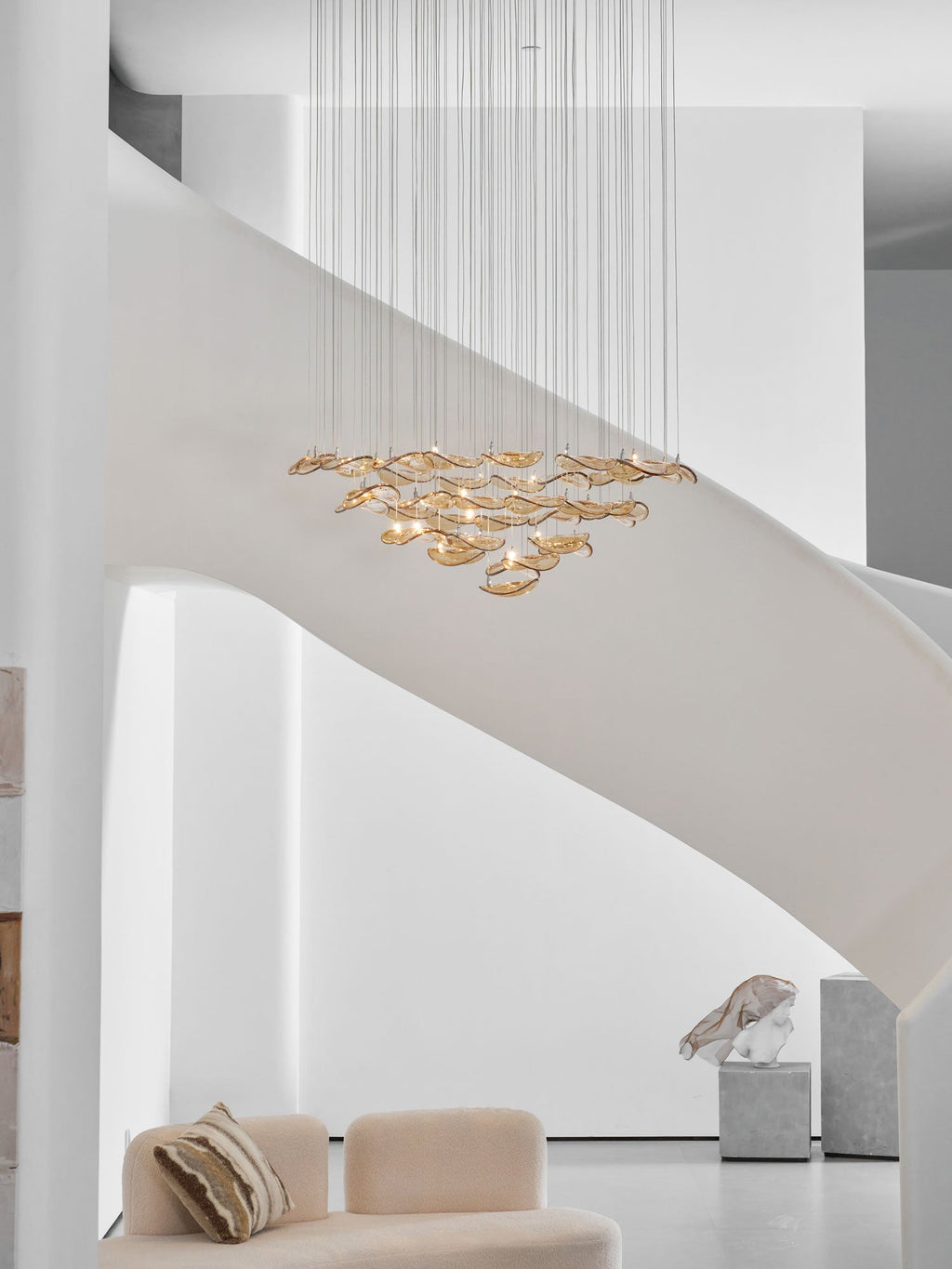 Bright white stairwell with a curved stair, where a Clérachic amber leaf pendant hangs well above a cream sofa.