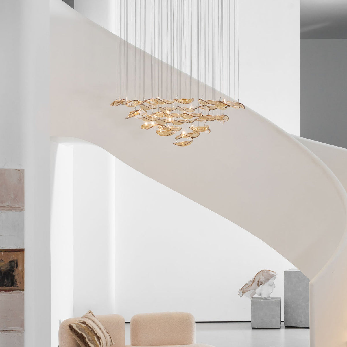 Pale stairwell with a Clérachic amber glass leaf pendant hanging in layers above a cream sofa and a stone plinth.
